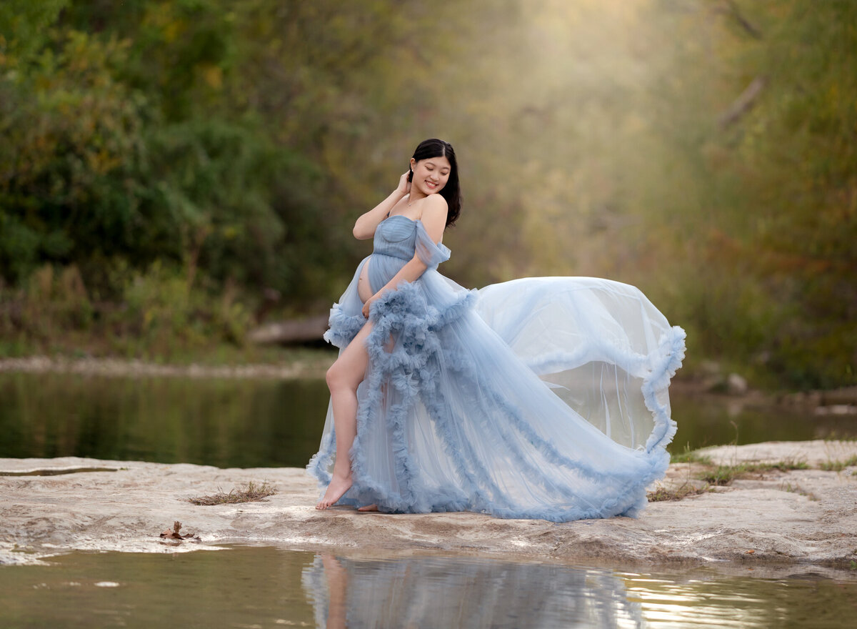 Maternity photo by a creek in Lakeway, TX featuring a mom-to-be in a flowing pastel blue gown