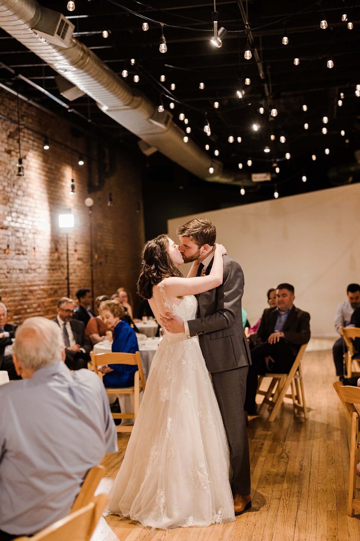 A bride and groom share a kiss during their first dance during their wedding reception in Plano, Texas. The bride is on the left and is wearing a long, flowing, intricate, white dress. The groom is on the right and is wearing a grey suit. Their guests watch on all around them from their reception tables.