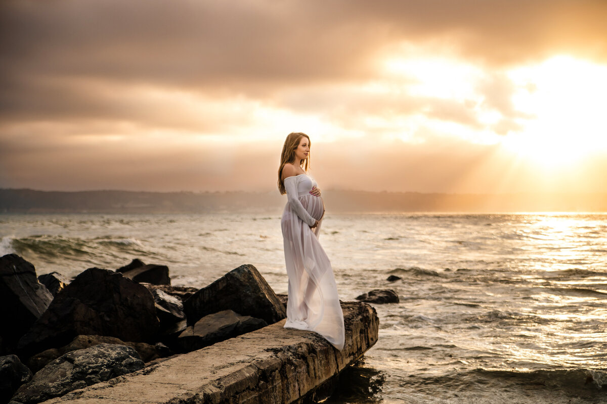 Maternity Photographer, an expectant mother stands on rocks near the ocean