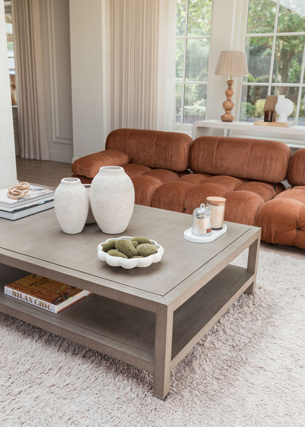 A cozy living room with large windows features a plush, rust orange tufted sofa, a light-colored rug, and a wooden coffee table adorned with decorative items and books. Two large, neutral lamps sit on a console table behind the sofa, and greenery is visible outside.