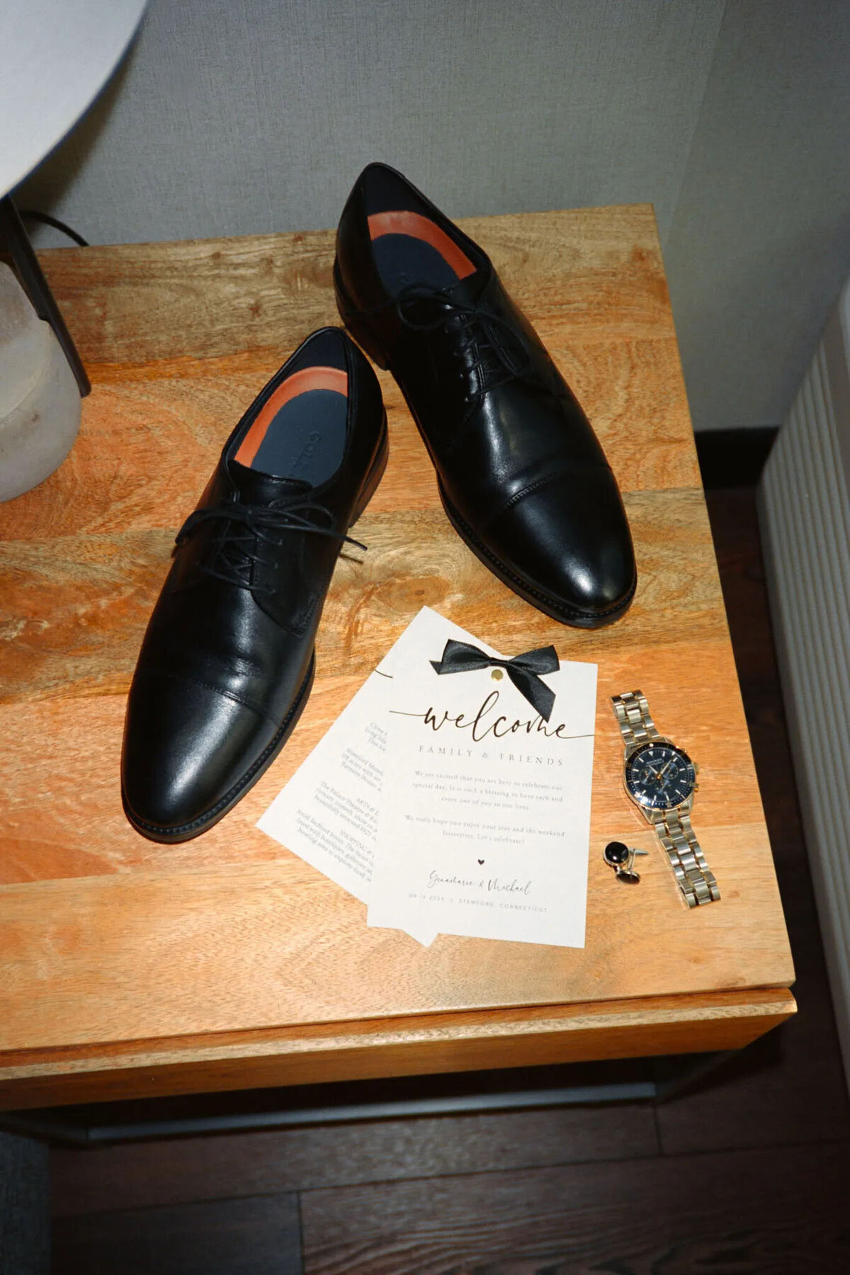 A pair of black dress shoes, a silver wristwatch, cufflinks, and a wedding welcome card with a bow tie illustration arranged on a wooden table, beautifully captured by a film photographer NJ.