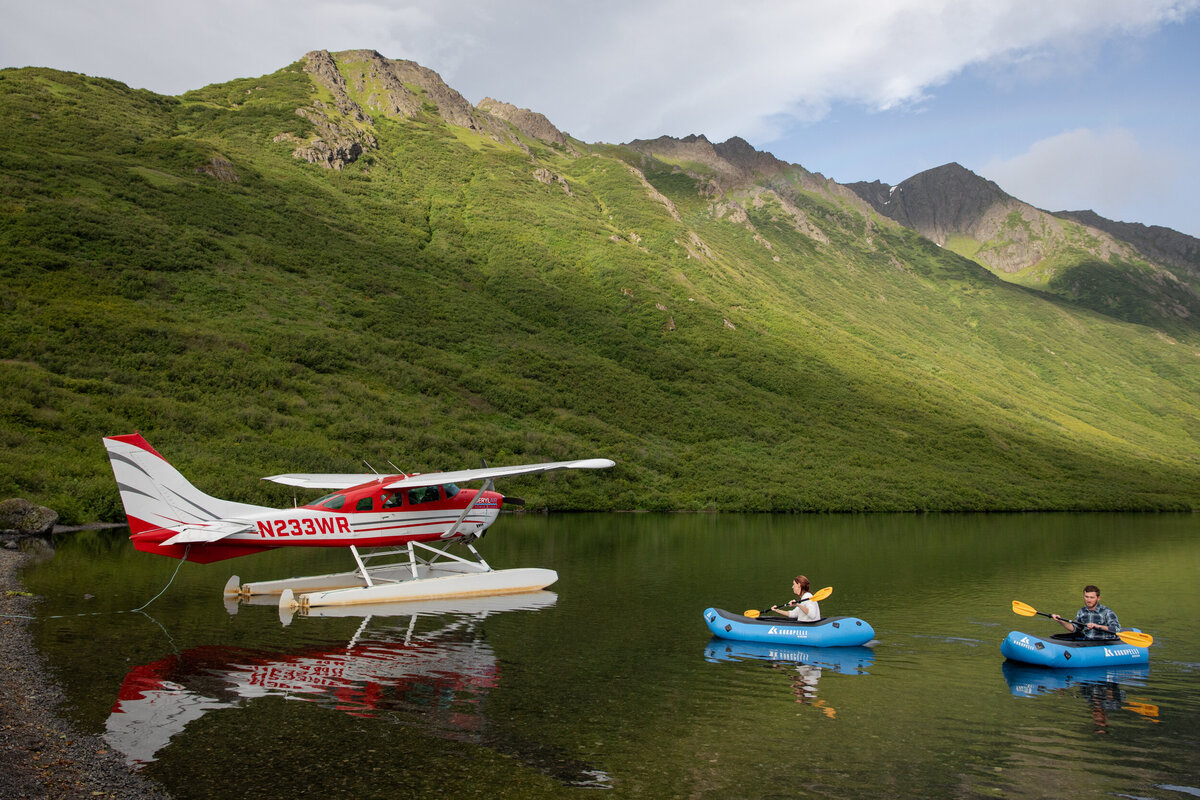 A bride and groom paddle next to a float plane in bright blue pack rafts in Alaska.