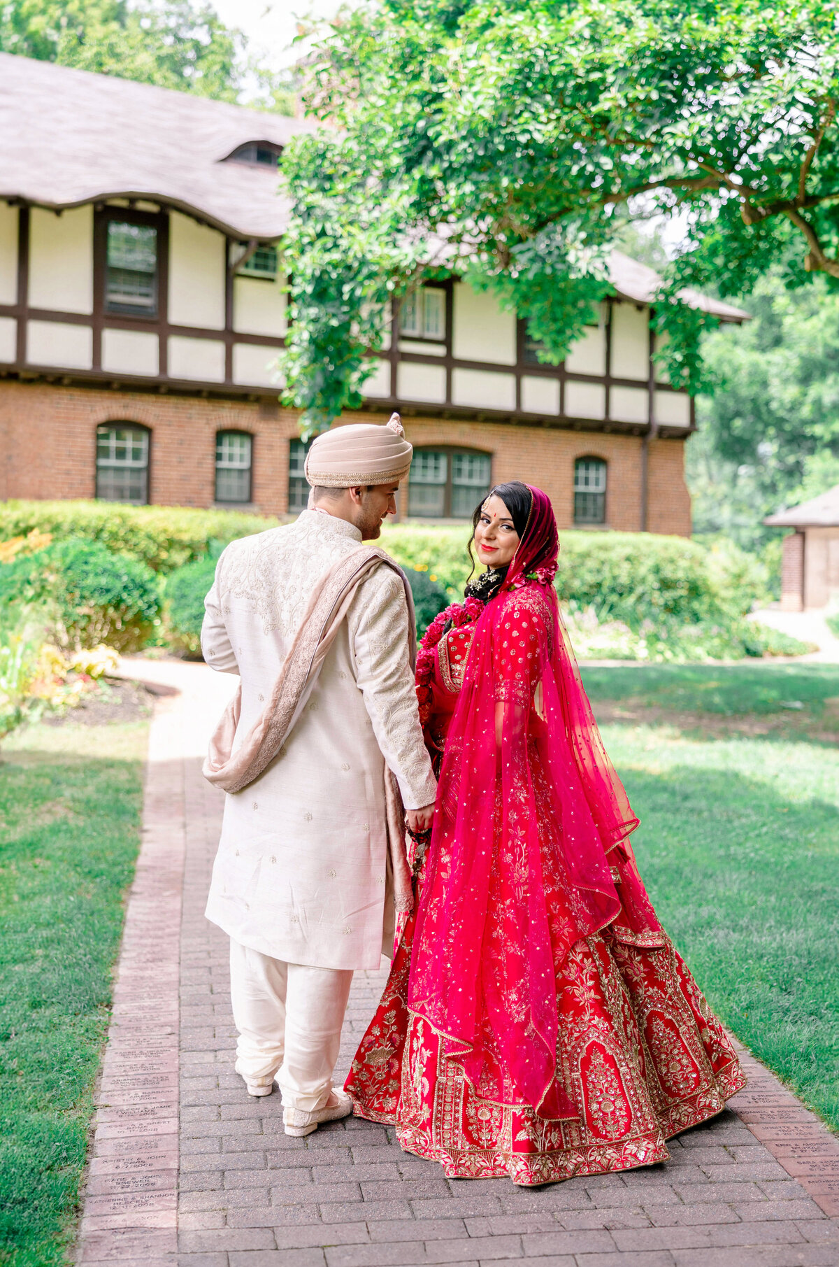 A couple dressed in traditional wedding attire stands on a brick path in a garden. The bride wears a red and gold embellished outfit, while the groom is in a cream-colored sherwani and turban. Greenery and a tudor-style building are in the background.