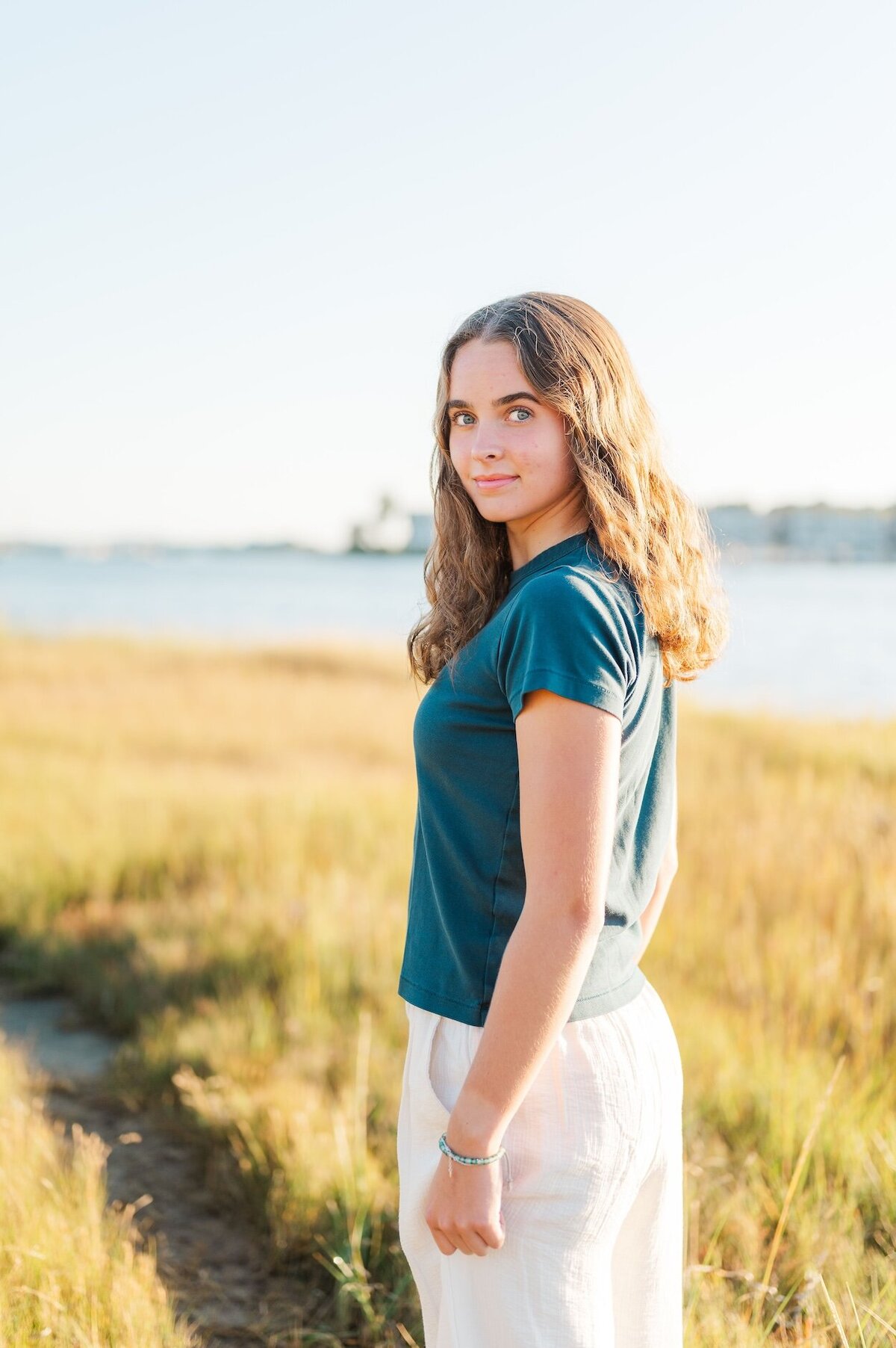A high school senior wearing a blue shirt and linen pants walks along a marshy path by the beach and peers over her shoulder.