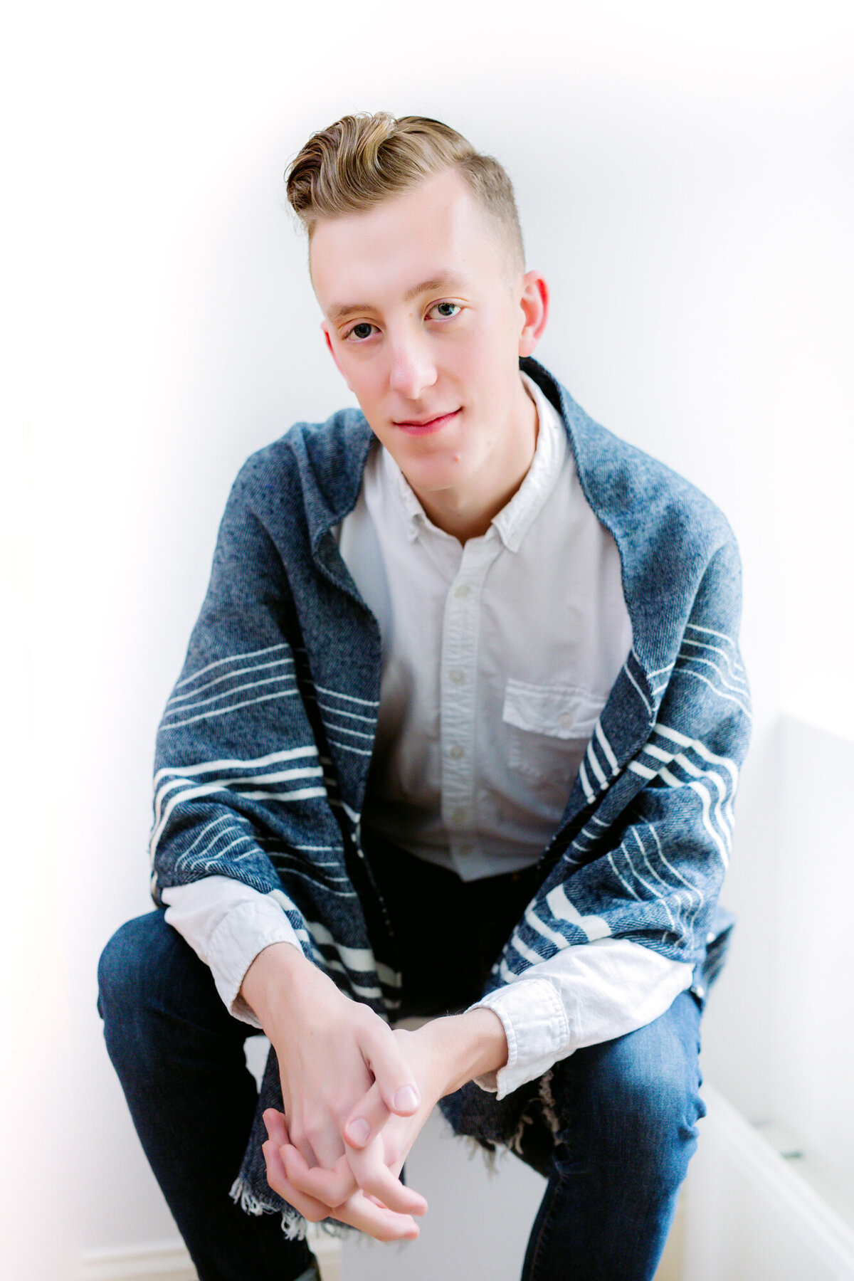 A young male adult headshot client wearing a shall over a button-down shirt seated with his hands in his lap