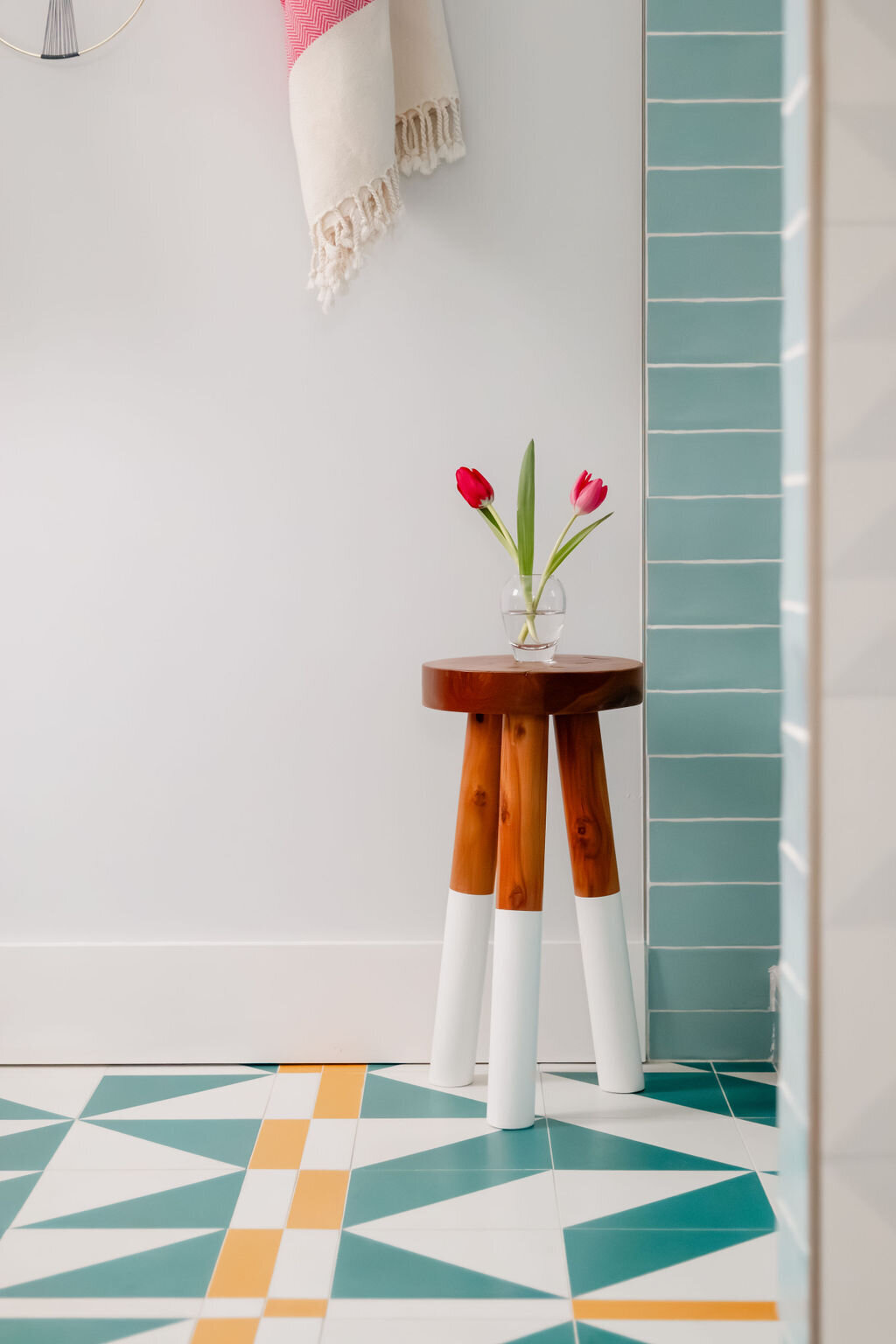 A brightly lit bathroom corner showcases a small, round wooden stool with white-painted leg bottoms. A clear glass bud vase holding two vibrant red tulips with green stems sits on the stool. Behind the stool, the wall is tiled with horizontal rows of light blue rectangular tiles with white grout. To the left, a white wall features a partially visible white and red patterned towel hanging from a hook. The floor is covered in a striking geometric tile pattern in shades of teal, white, and orange. A glimpse of a shower area with diagonally patterned white and light gray tiles is visible on the right.
