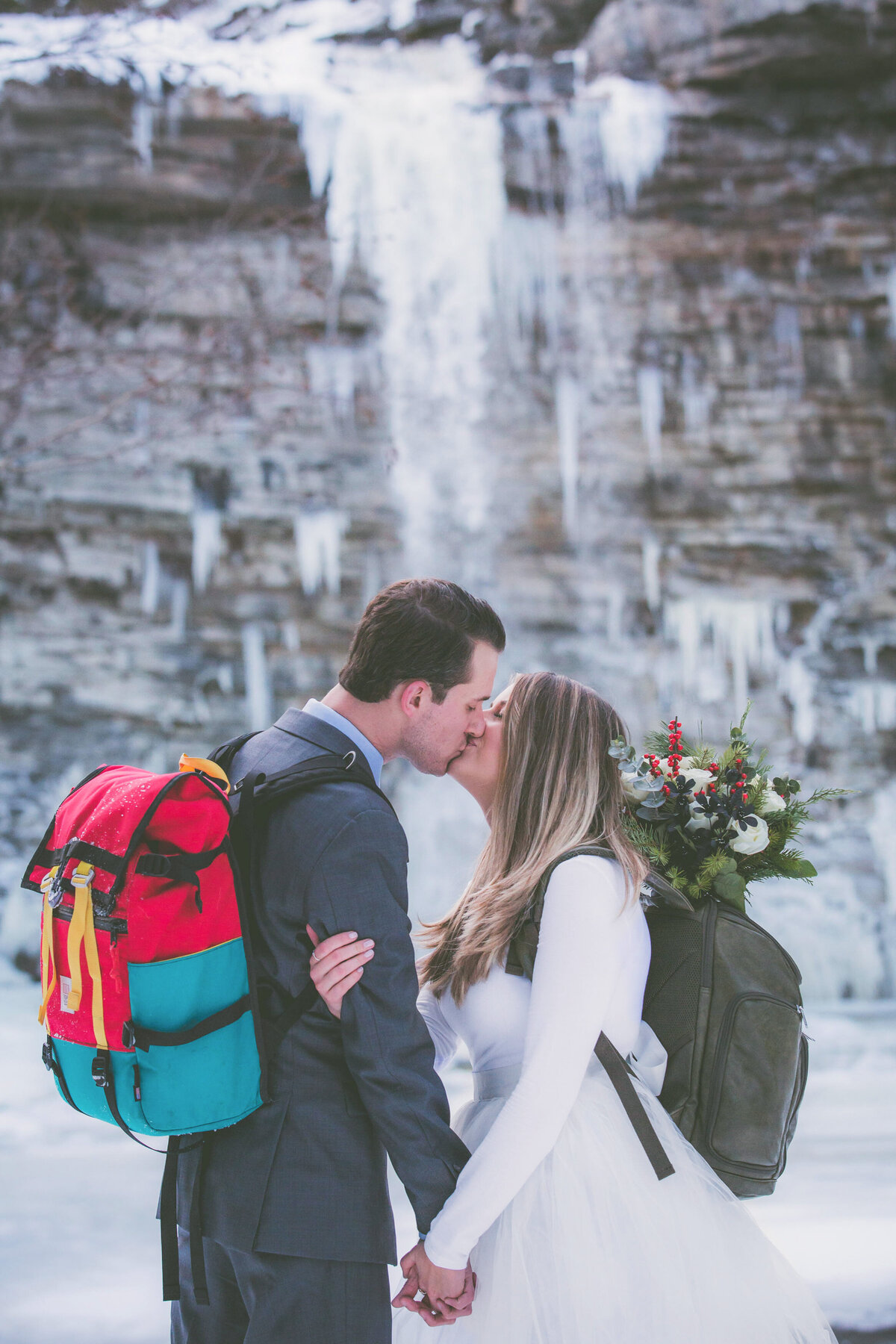 Bride and groom kiss with backpacks on with frozen waterfall behind them.