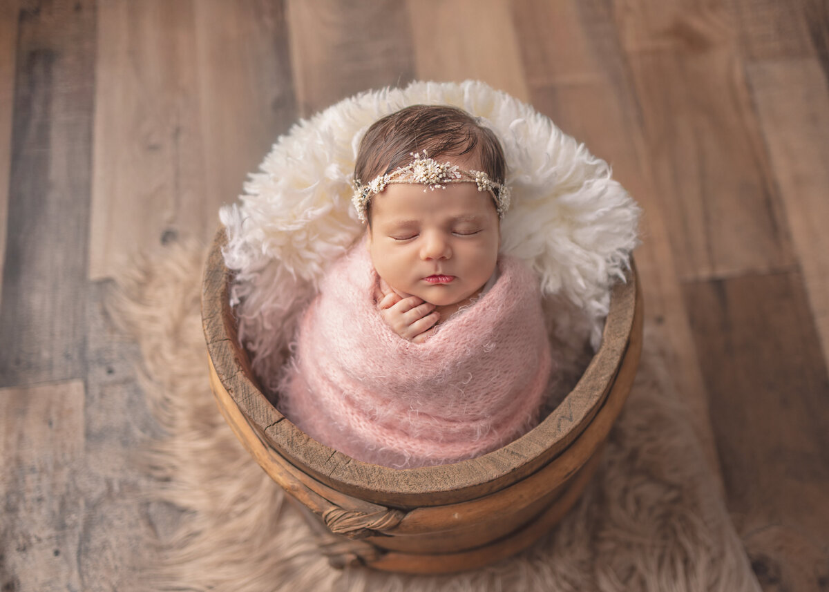 Newborn baby girl swaddled in soft pink wrap sleeping in a wooden bucket during a studio photoshoot in Bergen County NJ