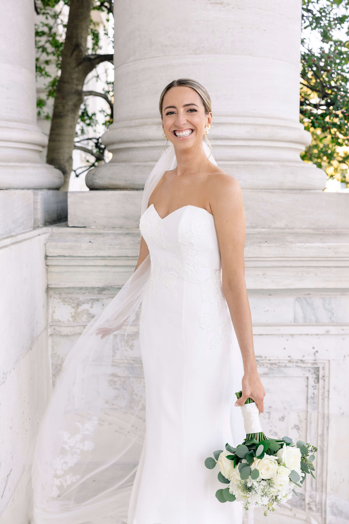 A smiling bride in a strapless white wedding dress holds a bouquet of white flowers and greenery. She stands near large white stone columns, with a delicate veil flowing in the breeze. Trees are visible in the background.