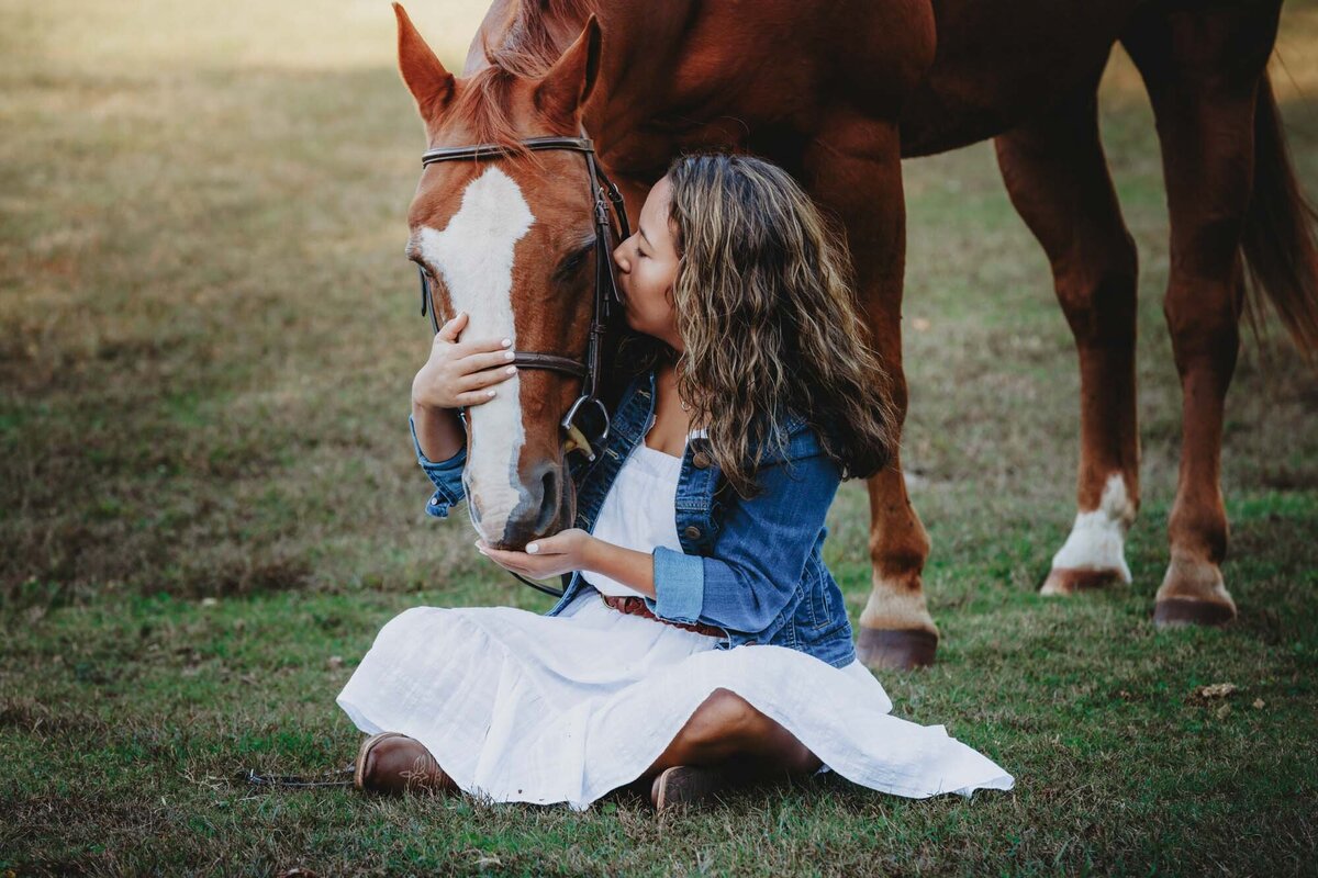 Girl in a white dress sitting on the grass while her horse kisses her hand.