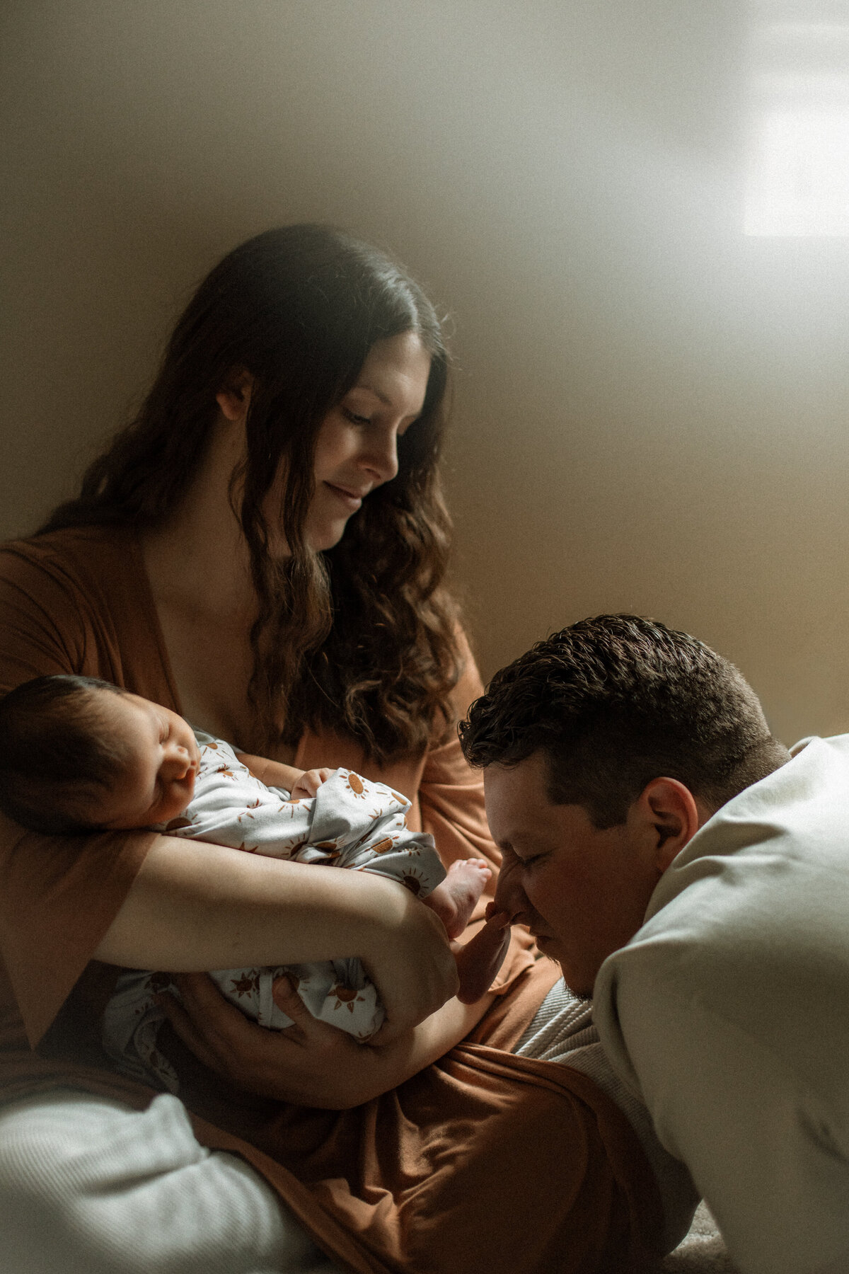 Mother cradling her newborn while the father gently kisses the baby’s feet, bathed in morning window light.