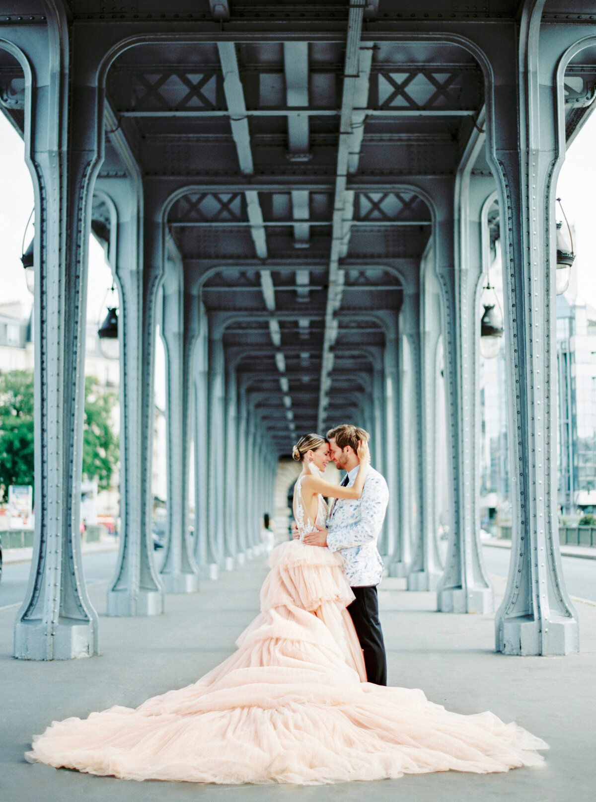 Couple during their Paris Engagement session under a bridge with lines of columns she is in a peach gown with a long train and he in black pants with white floral print jacket photographed by Italy wedding photographer