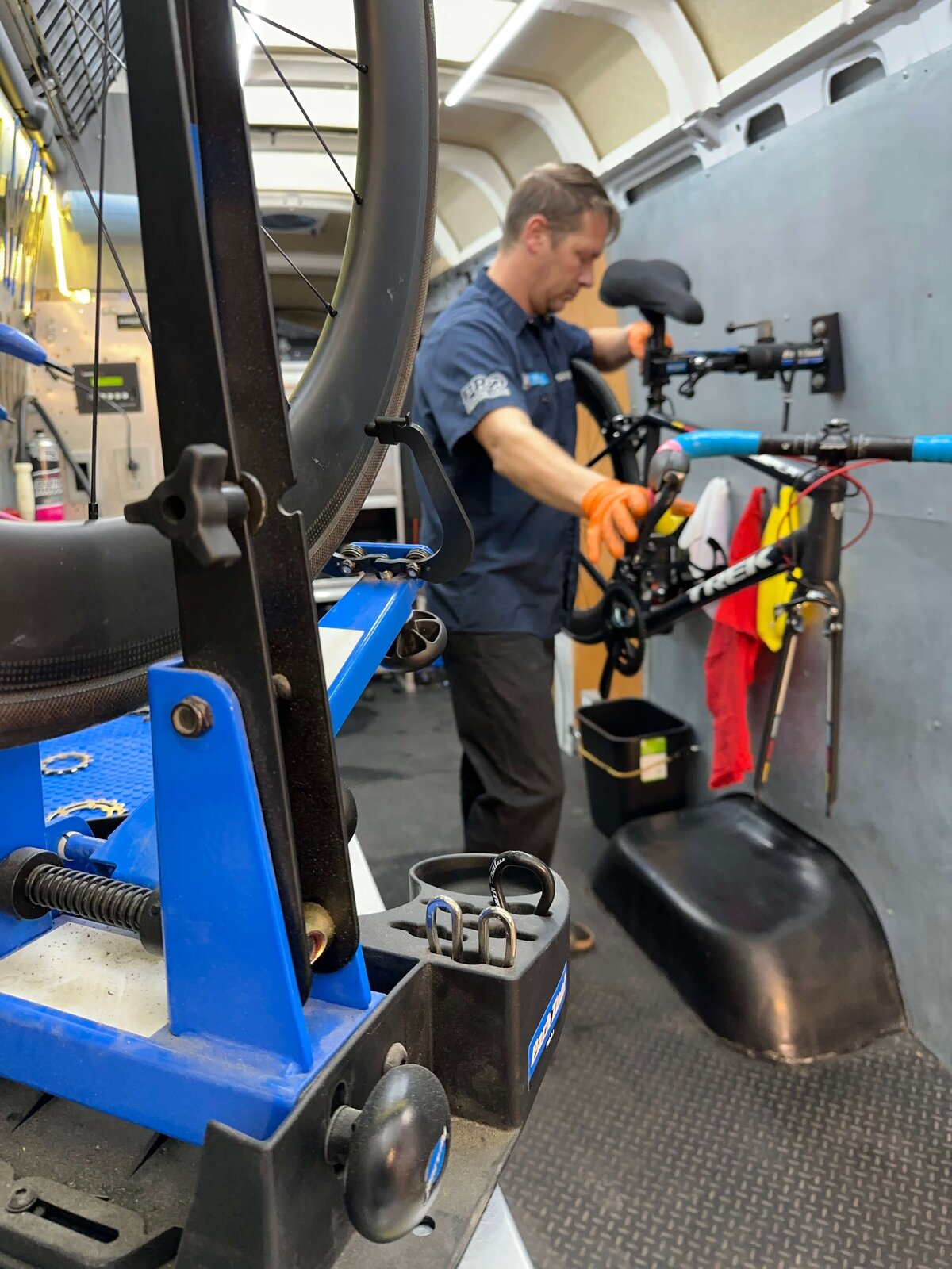 A mechanic wearing gloves is repairing a bicycle inside a workshop van. He is focused on adjusting the bike, with various tools and equipment visible around him. A bike stand is in the foreground, and towels hang nearby