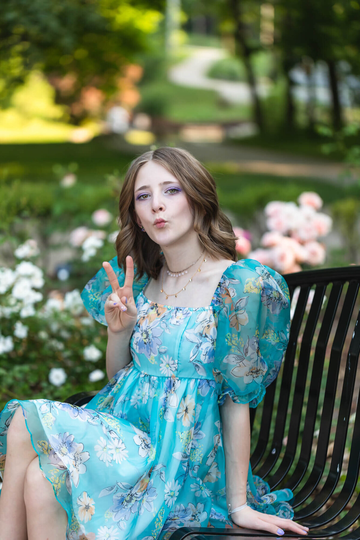 Fun portrait of a young teen girl in a floral blue dress throwing up the peace sign in a flower garden. Captured by Springfield, MO teen photographer Dynae Levingston.