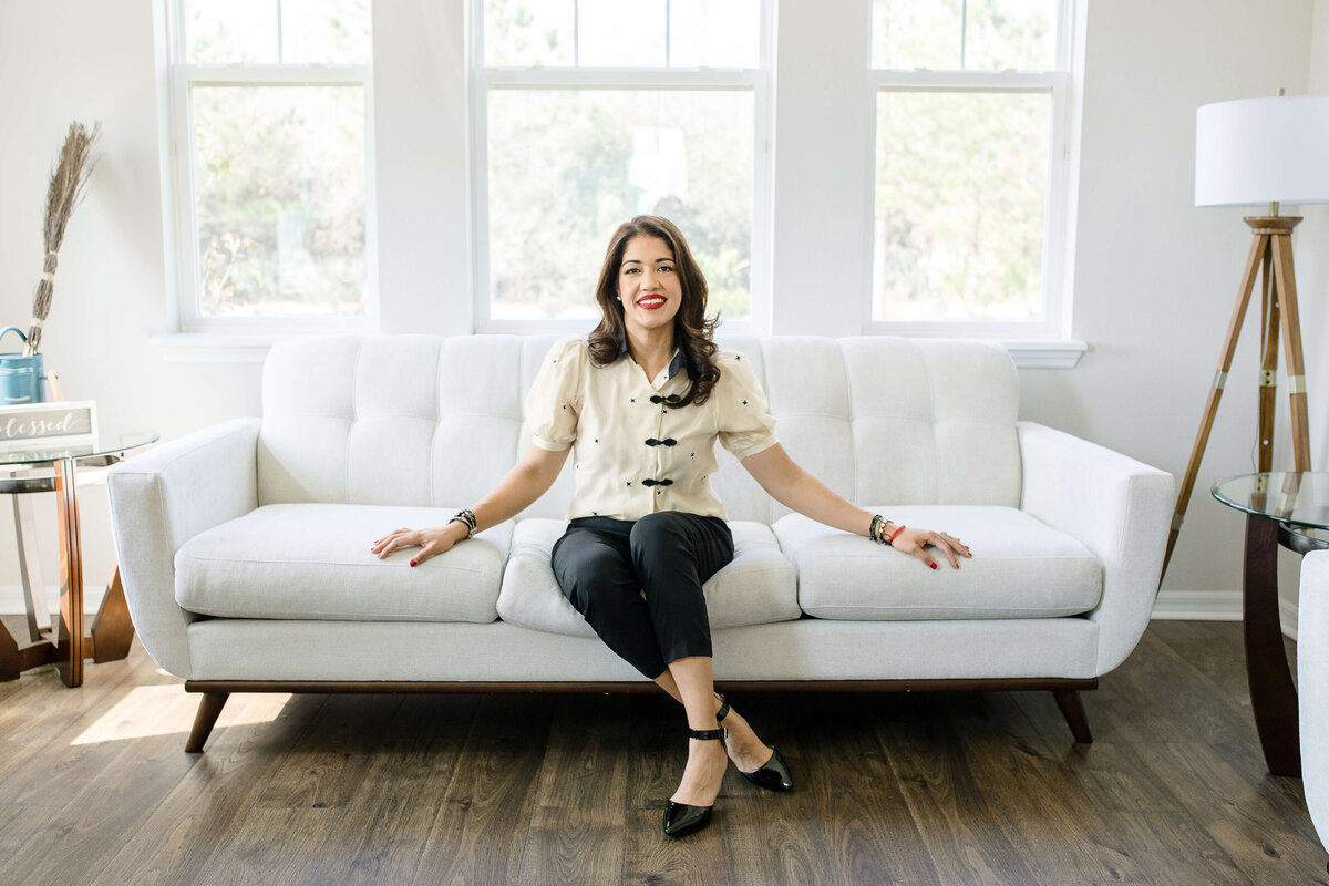 brand photographer near me photographs branding photo shoot ideas with woman sitting on a white couch in the living room with large open windows behind her bring in natural light for Orlando brand photo