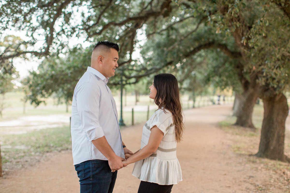 A couple stands, facing each other and holding hands with oak trees in the background