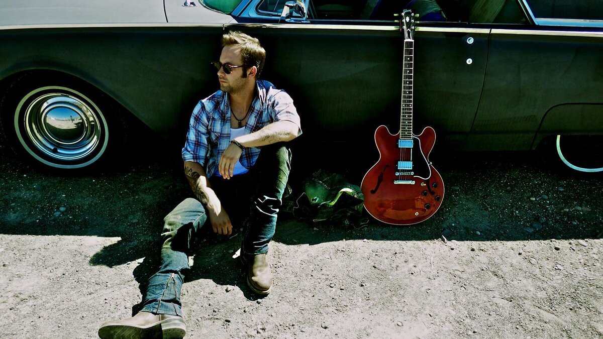 Country Music Artist Portrait Dallas Smith sitting on ground next to green vintage car red electric guitar leaning against car door beside him Roadshow Gallery