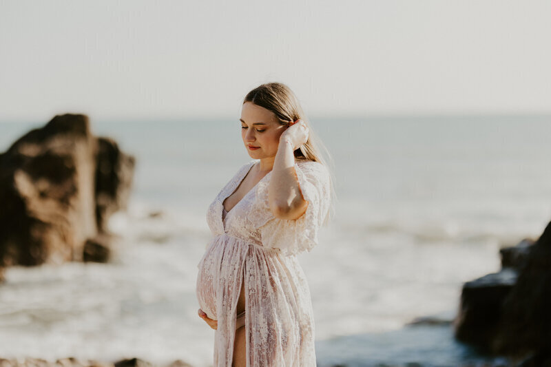 Femme enceinte en robe de dentelle blanche se passant la main dans les cheveux devant un décor de mer et de rochers lors d'un shooting photo en Vendée.