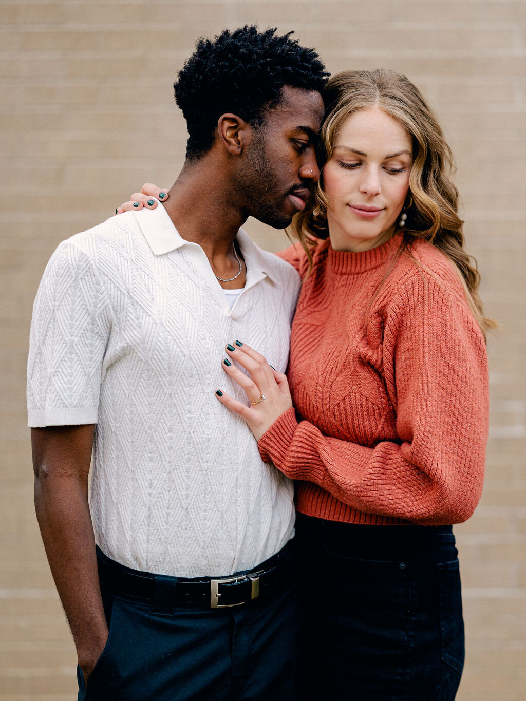 A couple stands against a white brick wall. They are embracing each other and not looking at the camera.