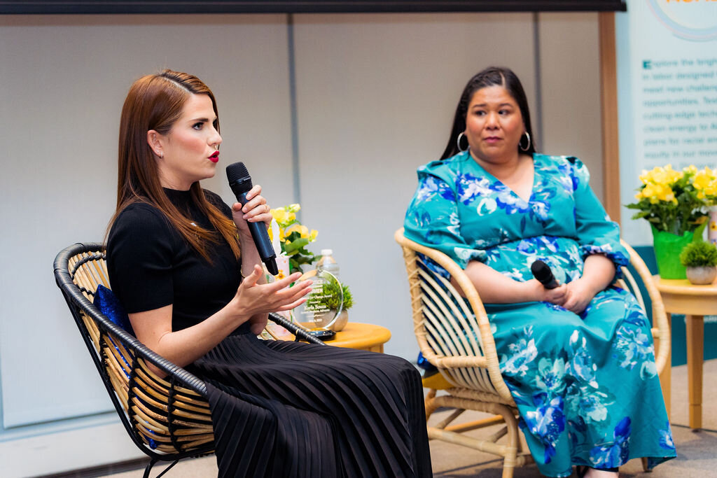 A woman speaks into a microphone while someone else sits in a chair next to her.