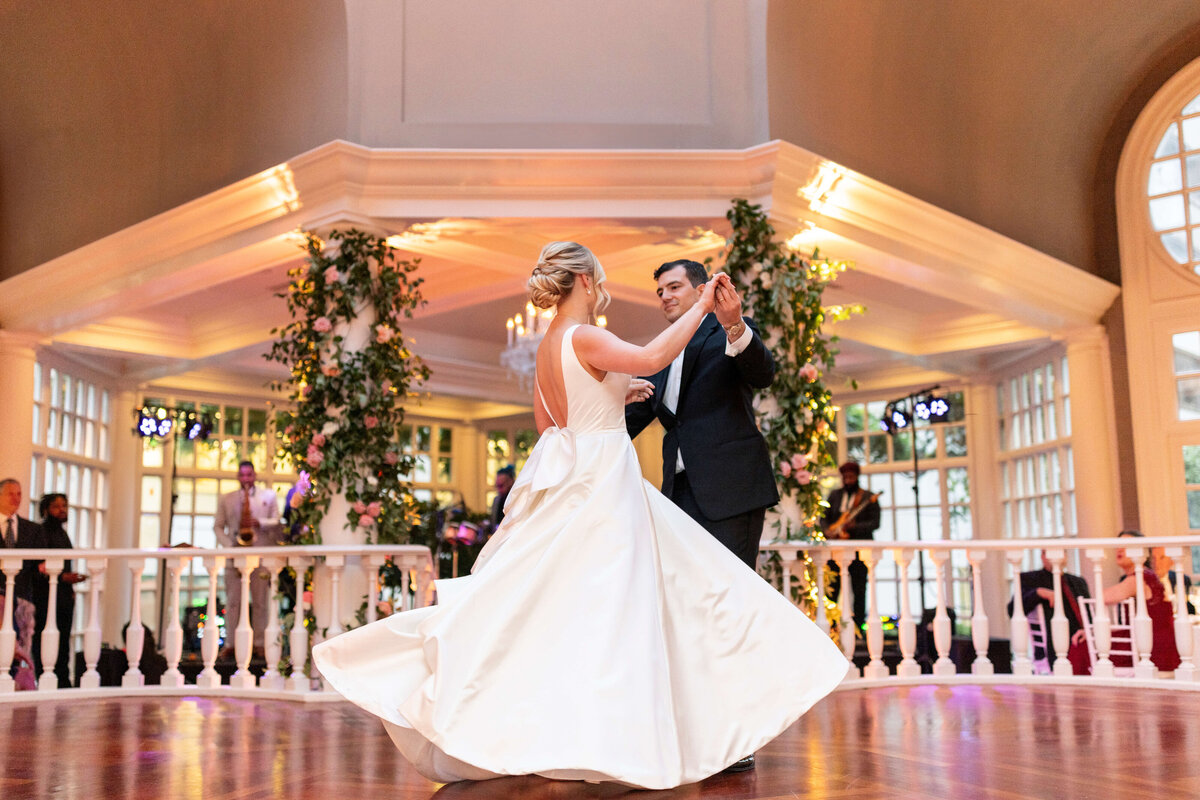 A bride and groom share their first dance in an elegant ballroom adorned with greenery and soft lighting. The brides white gown twirls gracefully as the couple dances. Musicians are visible in the background.
