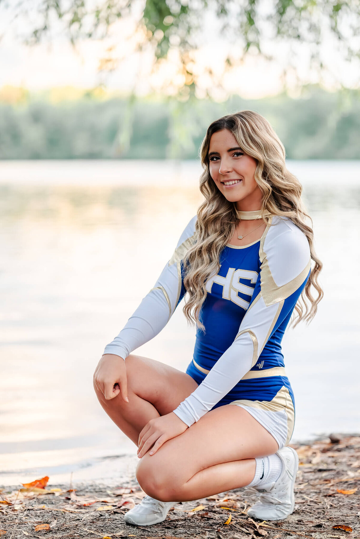 A high school senior, wearing her blue, gold, and white cheer uniform, kneels down in front of a lake. Photo by Justine Renee Photography in Chesapeake, Virginia.