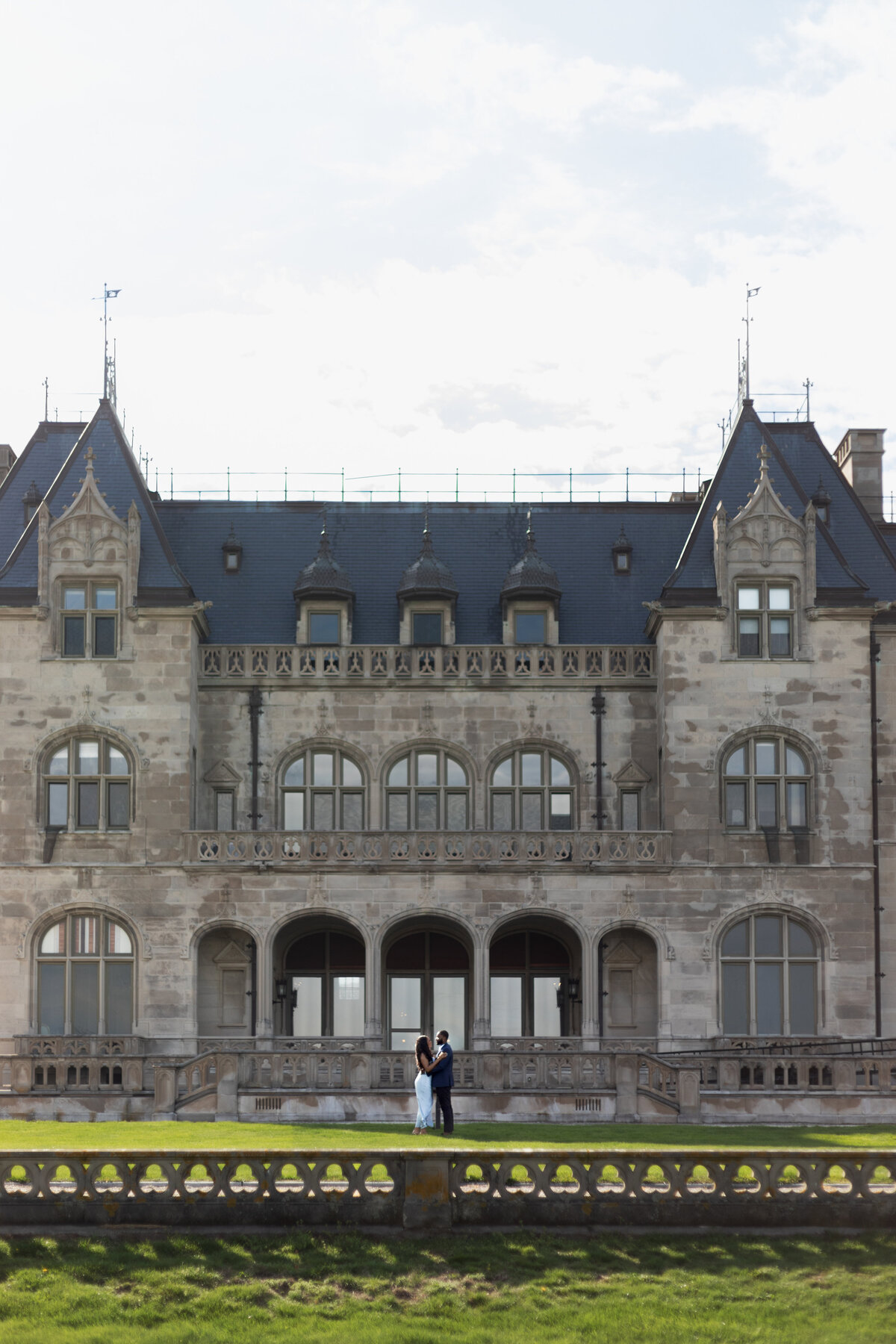 Ochre Court Mansion  | Kelsey Sheehan Photography Timeless Rhode Island Weddings | A couple stands closely on a balcony, framed by a grand stone mansion with arched windows and intricate details. Overcast sky hints at a calm, historic ambiance.