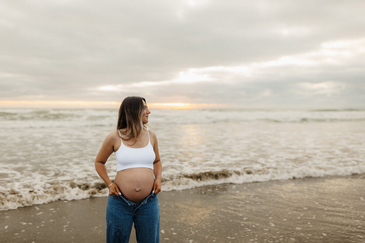 A pregnant woman standing on the beach in a white top and jeans while she's photographed by christchurch photographer lucy jane