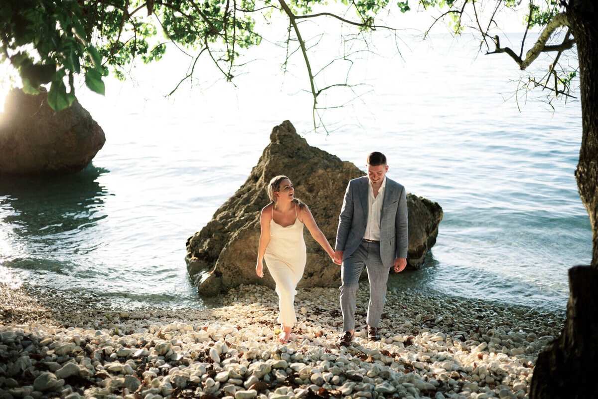 The engaged couple is holding hands while walking on a rocky seashore in Round Hill Hotel and Villas, Jamaica.