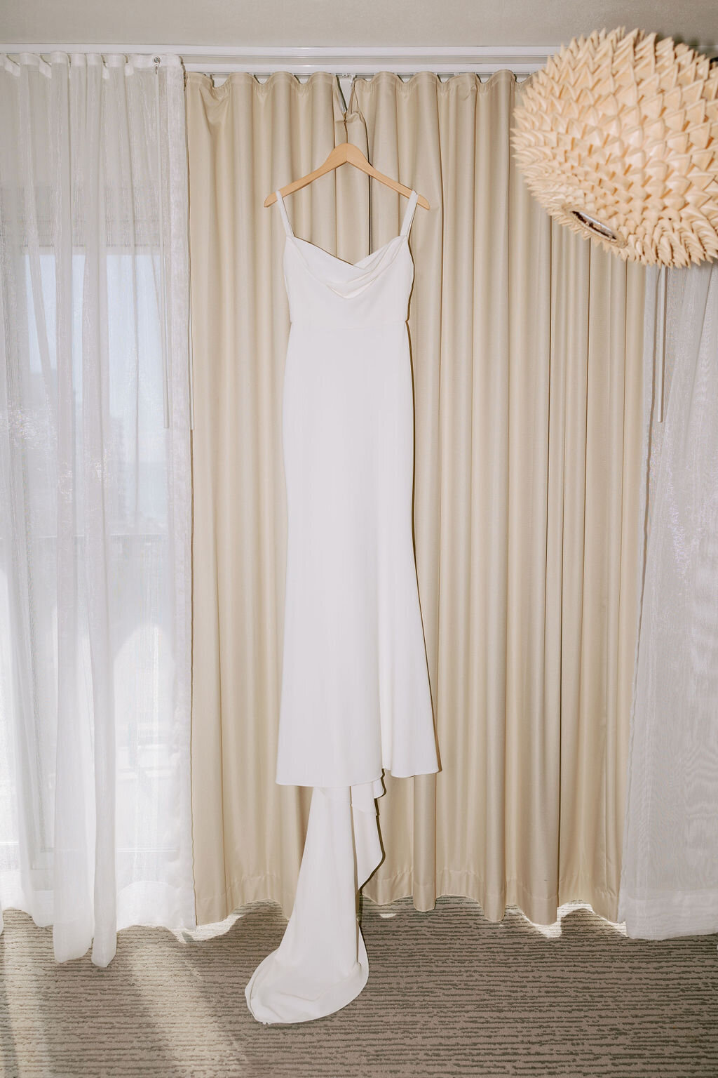 bride modern crepe wedding dress hanging in laylow waikiki hotel room in front of tan curtains