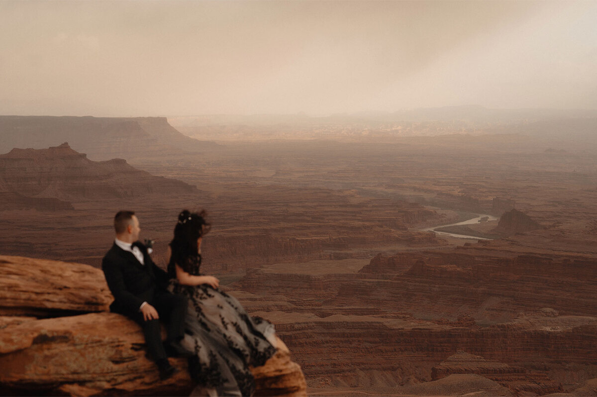 bride and groom sit on the edge of a rock as they look out at the beautiful scenery at Dead Horse Point State Park in Moab, Utah
