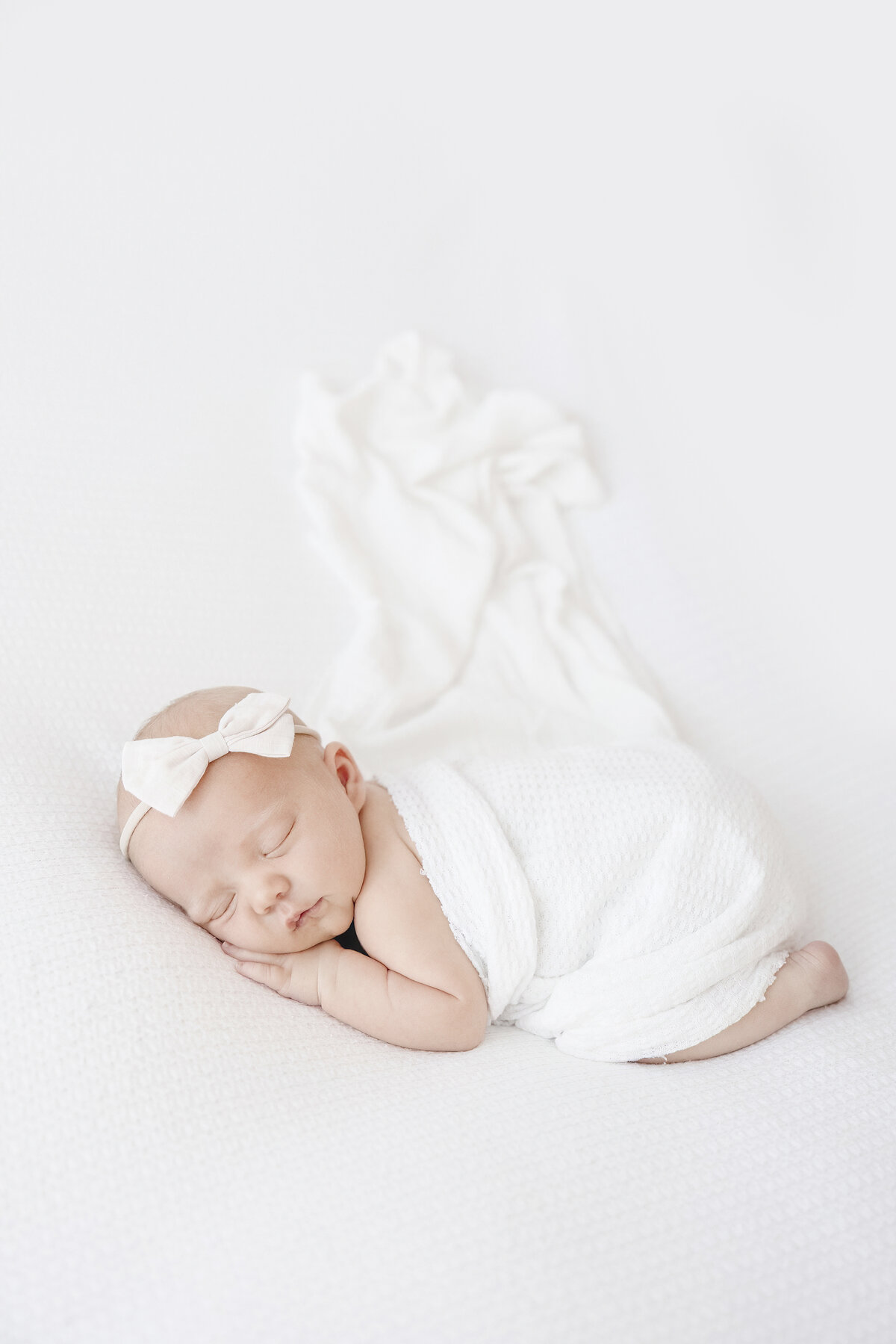 Newborn baby girl sleeping on belly with white wrap and white bow