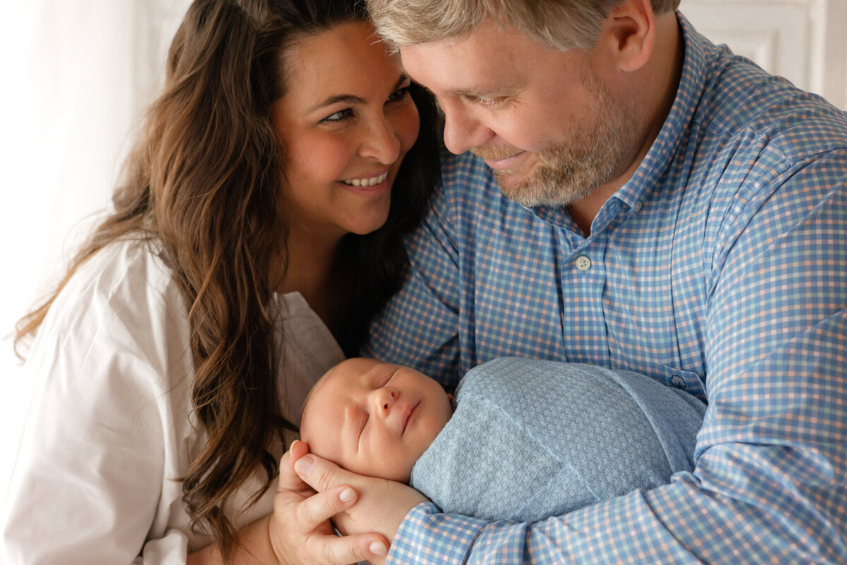 Mom and dad looking at each other while holding their baby boy wrapped in a light blue wrap