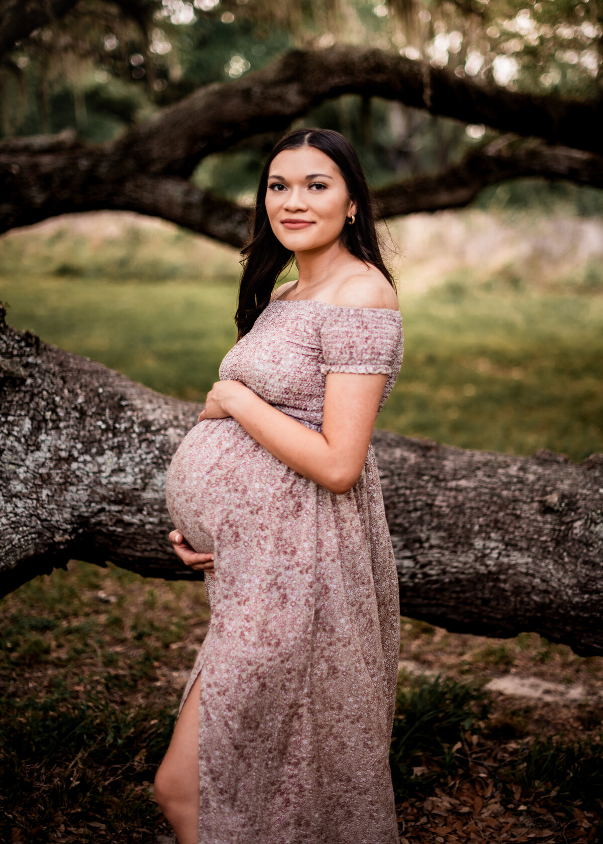 A pregnant woman stands sideways and holds her bump while she smiles at the camera.