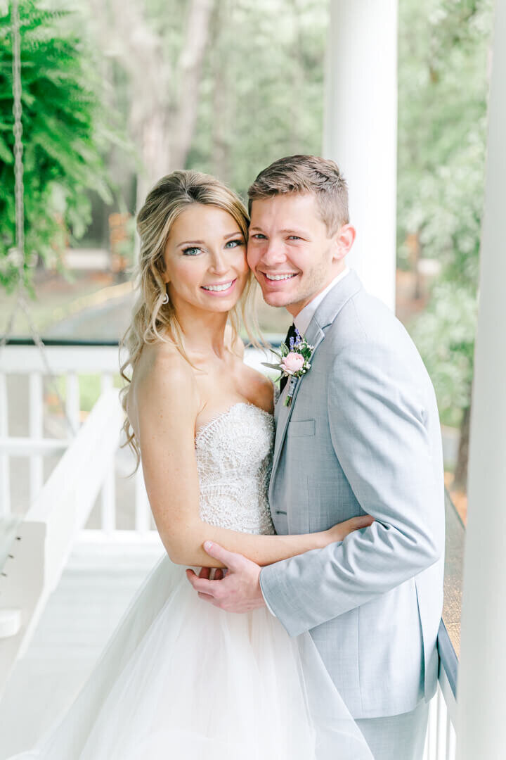 A bride and groom in a grey suit hug cheek to cheek on a porch for their Arkansas wedding photographer