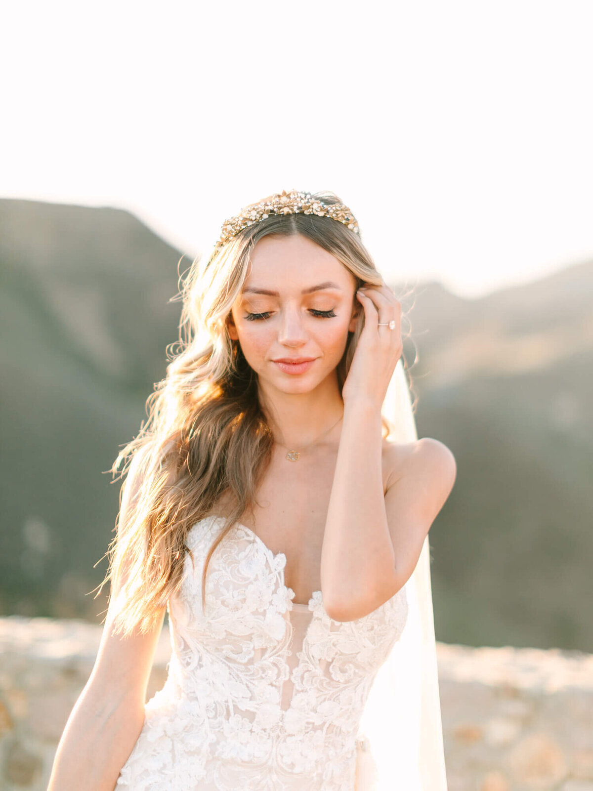 Bride in lace wedding dress and golden tiara poses outdoors, sunlight highlighting her flowing hair against a blurred mountain backdrop