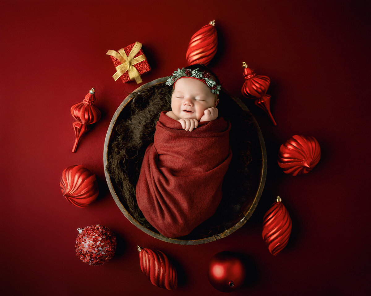 A smiling baby girl sleeps in a wooden bowl surrounded by red christmas ornaments