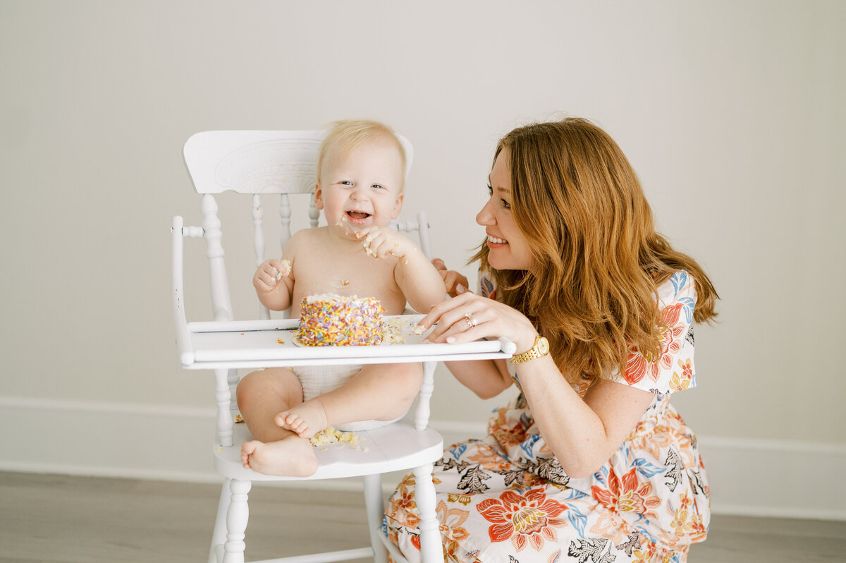 One year old baby boy sits in white high chair eating cake with mom smiling at him
