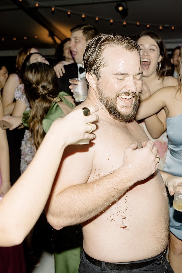 Groom with his shirt off at wedding reception while people pour drinks on him