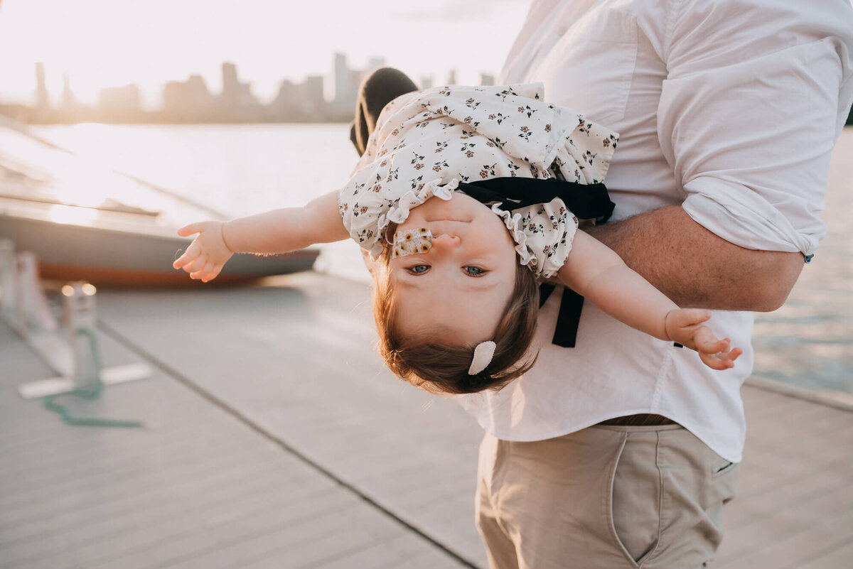 A father holds his daughter upside down , she looks directly at the camera with her arms out wide at her sides