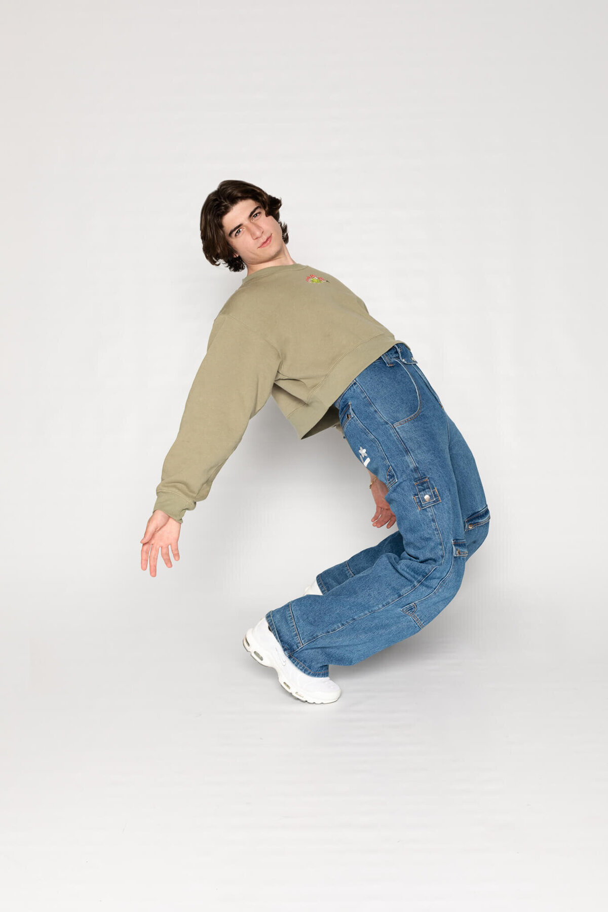 A person with dark hair is wearing a beige sweater, blue jeans, and white sneakers. They are striking a dynamic pose, leaning back with arms extended to the sides, against a plain white background.