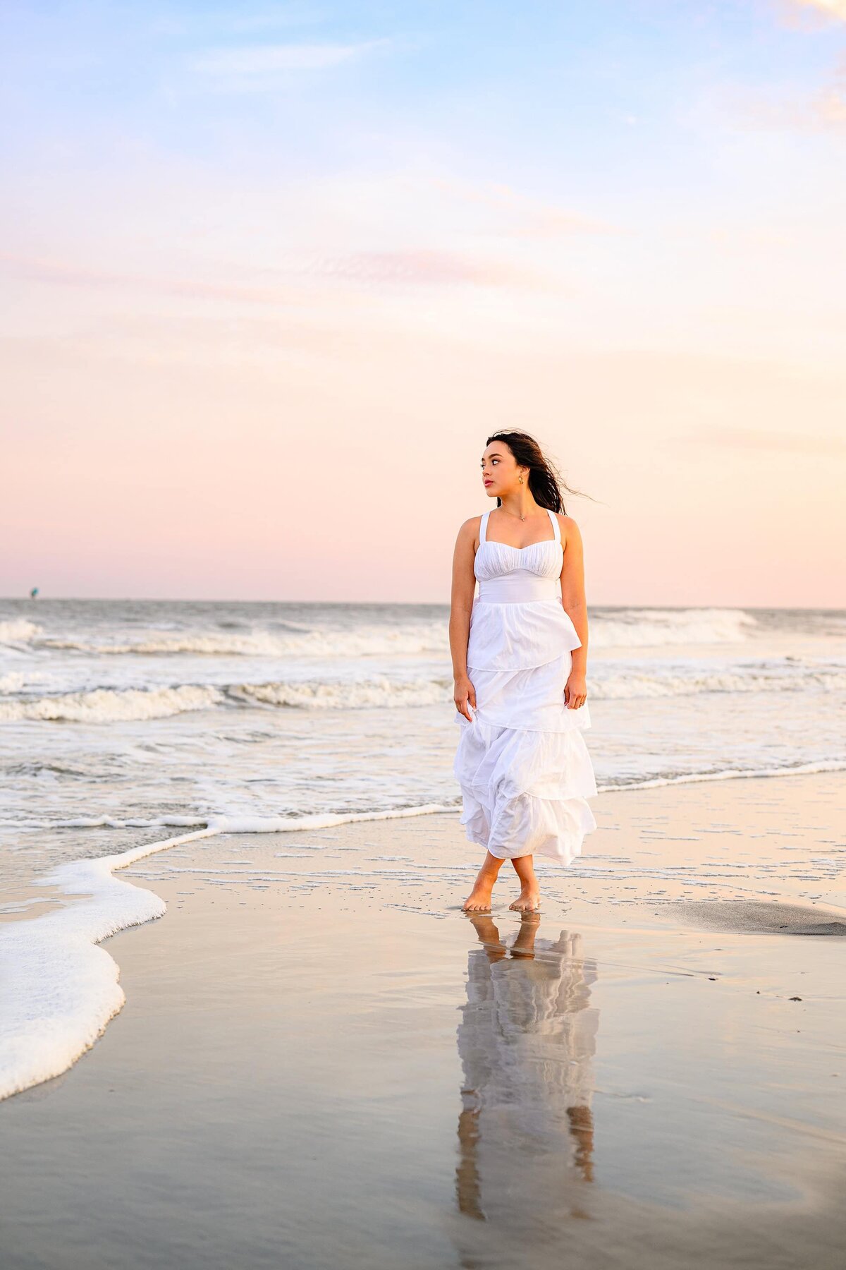 charlotte senior stands on beach wearing a white dress with water reflection in front of her on a beach