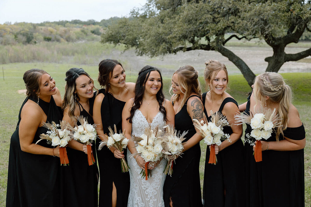 A radiant bride surrounded by her bridesmaids in elegant black dresses holding pampas and ivory rose bouquets. This rustic outdoor wedding captured natural glam bridal makeup and effortless waves styled by Mistique Makeup. Perfect for brides searching for wedding day hair and makeup in Dallas, San Antonio or Austin, or looking for a mobile bridal hair and makeup team.