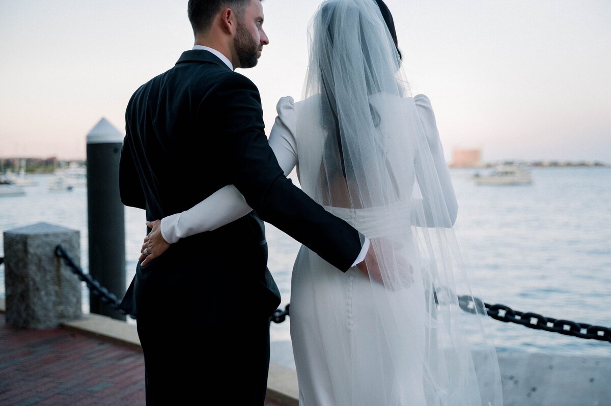 a bride and groom with their arms around each other standing near the Boston Harbor