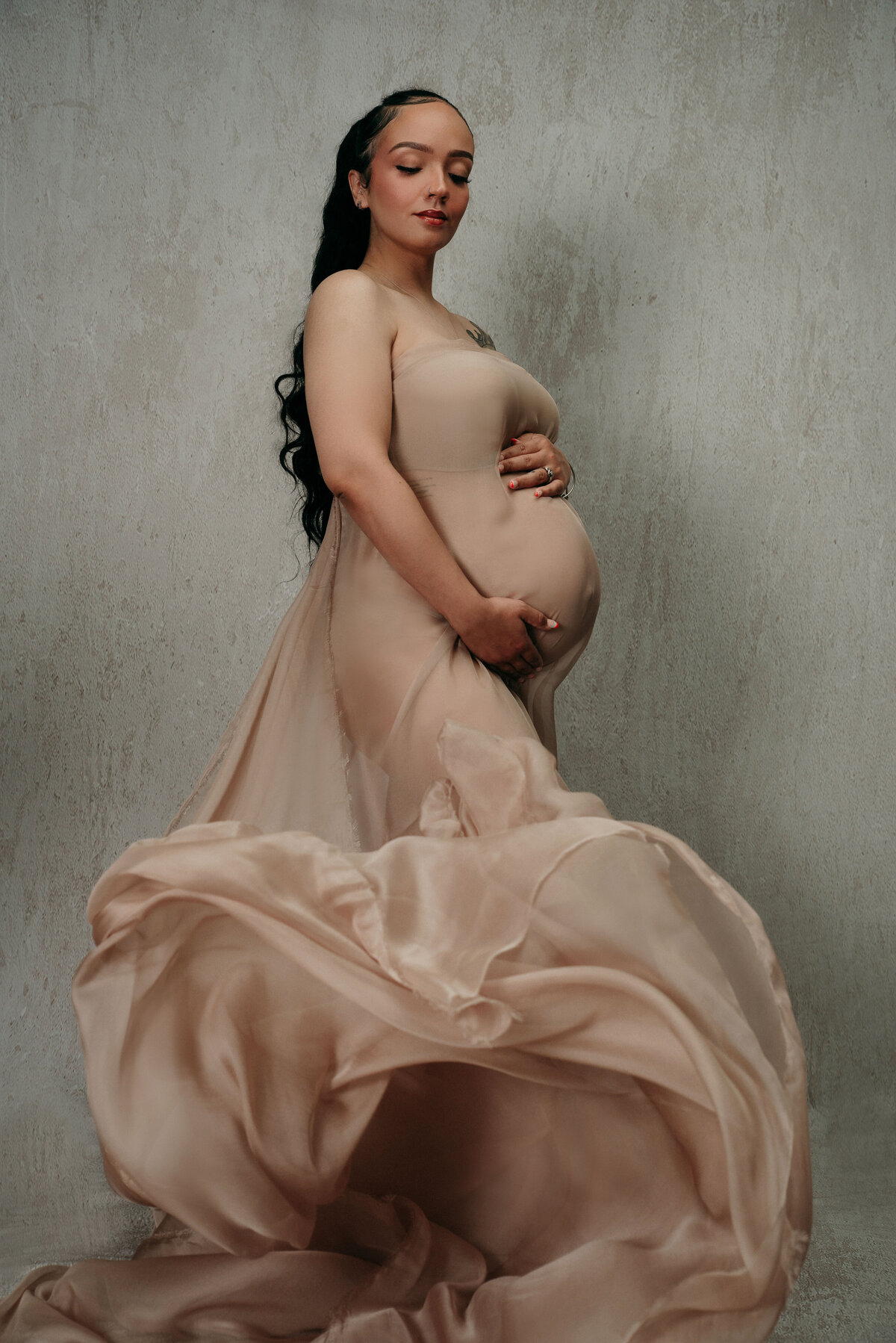Pregnant woman with long black ponytail posing holding baby bump wrapped in tan chiffon fabric flowing around her