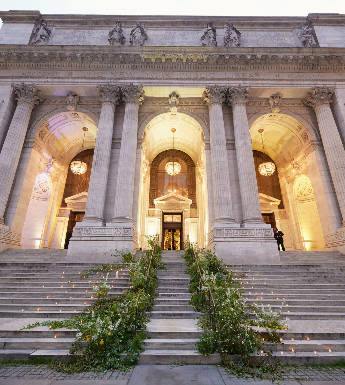This is where the photo was taken at New York City, The New York Public Library by photographer Shawn Connell. The grand stone entrance features wide steps leading to large arched doorways adorned with elaborate carvings and statues. Flowers and plants line the staircase, and warm lights illuminate the building, showcasing the neoclassical architecture with tall columns and intricate details. Videographers: Aaron Navak Films