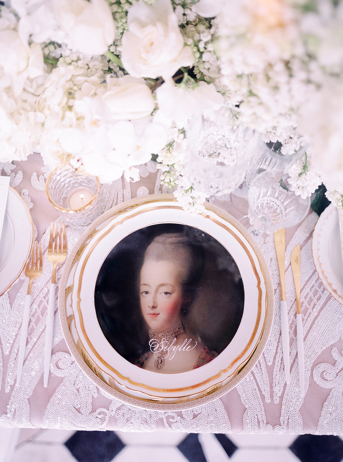 The photo was taken at Paris, France - Chateau Vaux le Vicomte, one of the top destination wedding venues in France, beautifully captured by the talented photographer Audrey. The scene showcases a decorative table setting, including a plate with a stunning portrait of a historical woman dressed in elegant 18th-century attire. Surrounding the portrait are exquisite gold-rimmed cutlery, a delicate white lace tablecloth, and elaborate floral arrangements featuring stunning white blooms. East West Films were the videographers capturing the beautiful moments at the event.