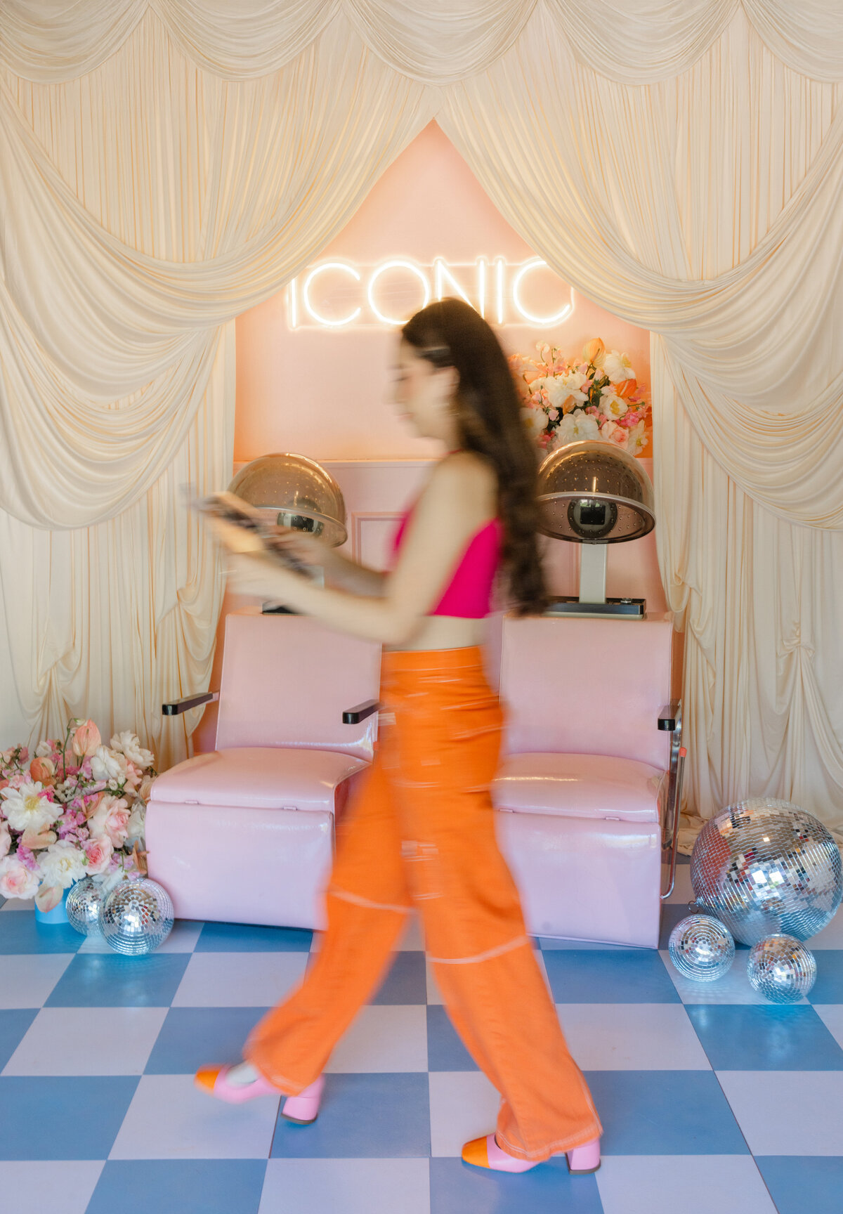 Blurred motion shot of a woman walking past a retro pink salon setup with disco balls and an "ICONIC" neon sign in Greenville, SC. Styled branding image by Dani Lacey.