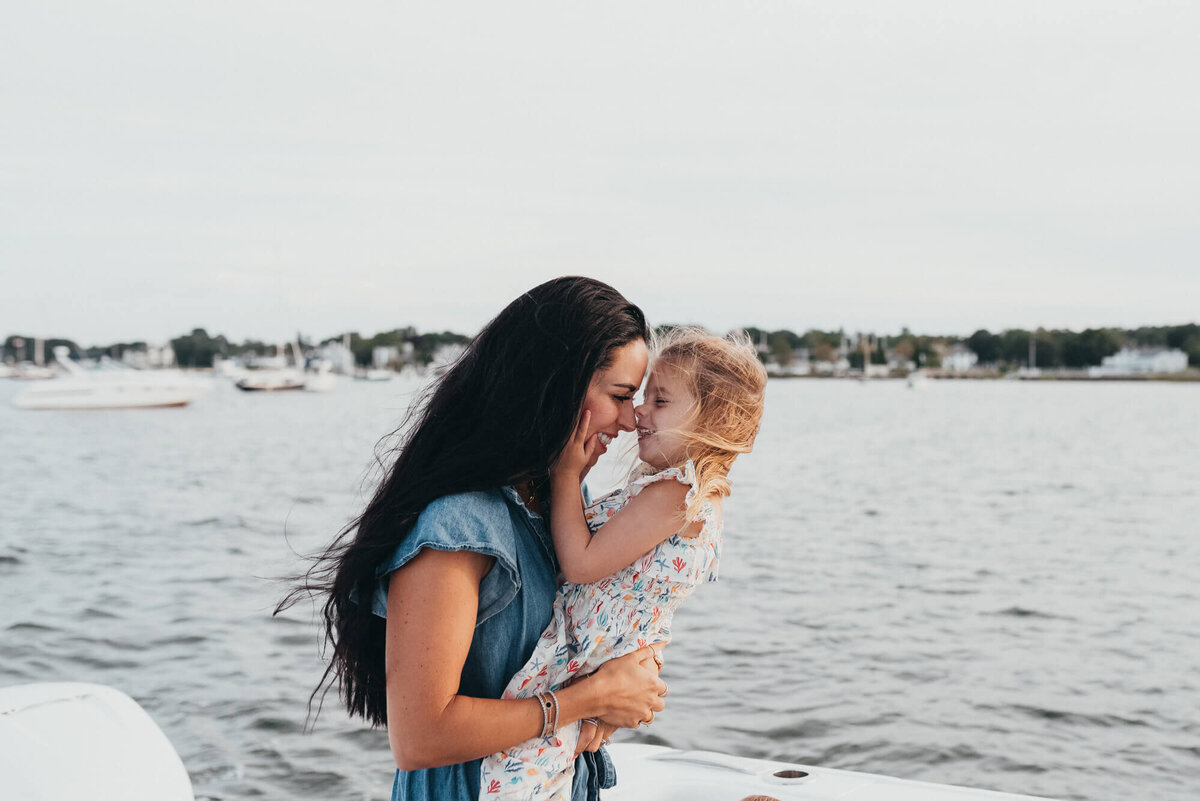 A young mother with long black hair stands next to the ocean with her daughter, her daughter holds her face and they touch noses during their photoshoot with Boston Family Photographer, Allison Wolf