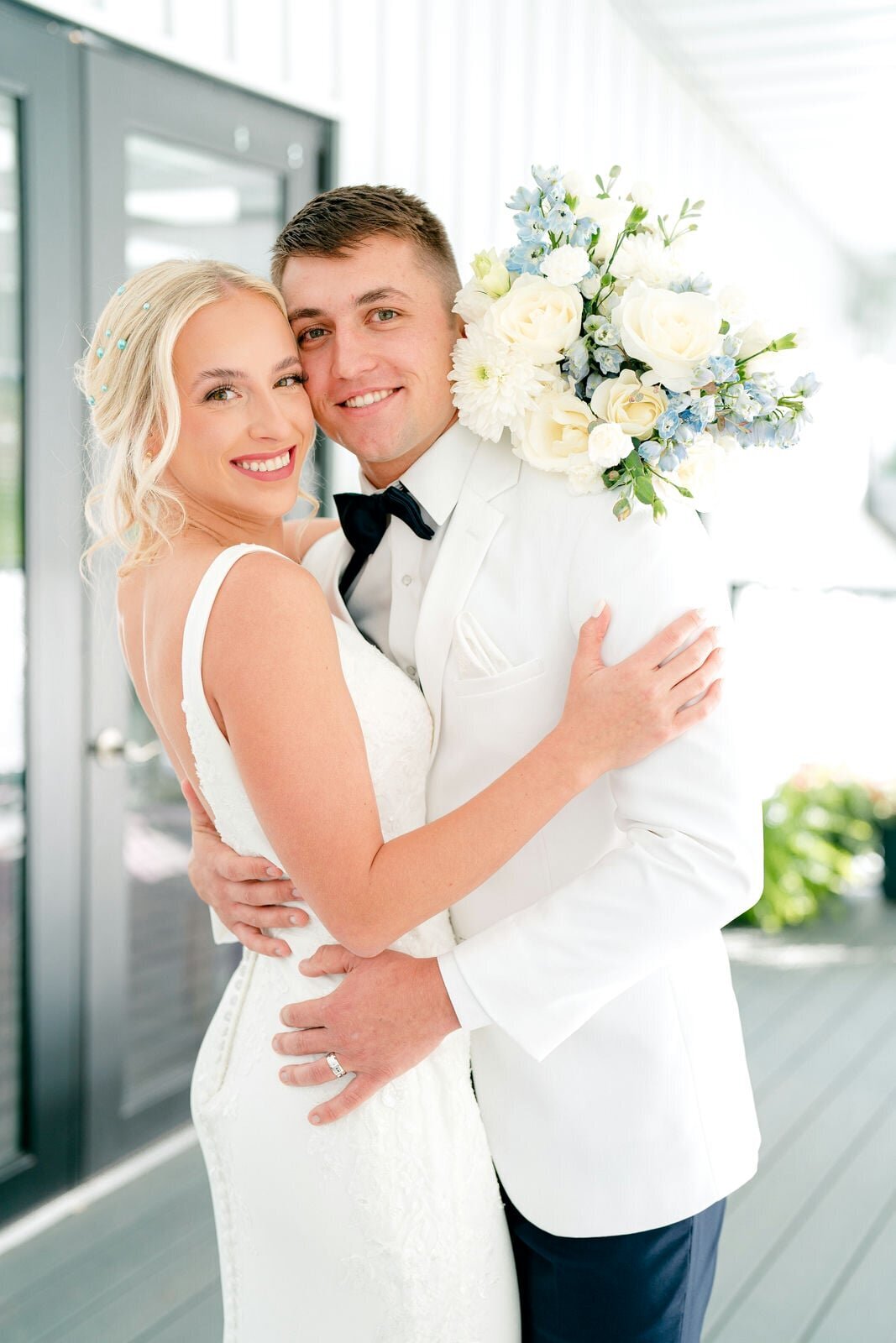 A close-up of a bride and groom smiling brightly on their wedding day. The bride wears a white gown, and the groom holds a bouquet of white and blue flowers over his shoulder, capturing their happiness and love.